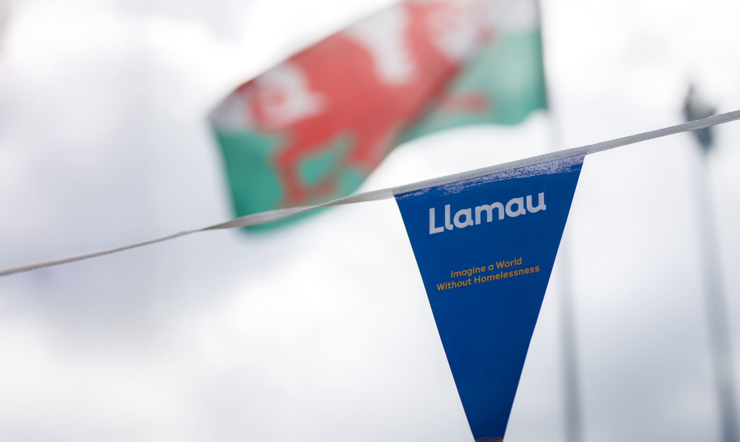 Small blue triangular flag with Llamau logo and strapline, with an out of focus Welsh flag in the background.