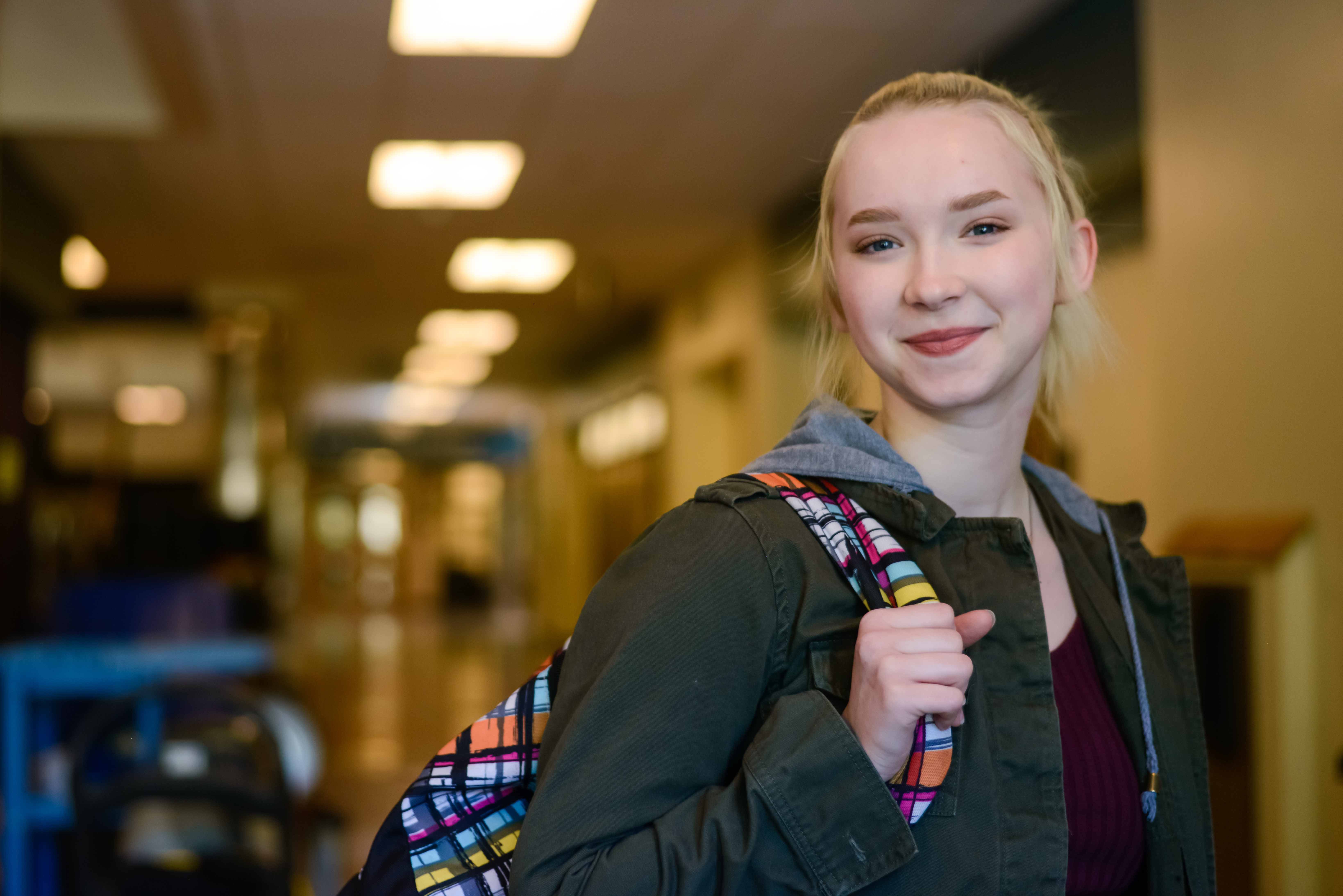 A girl in a school corridor, holding on to her backpack and smiling.